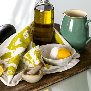 Table setting with Veggie tea towel, an olive oil bottle, a lemon, a ceramic bowl, and a green pitcher on a wooden borad.