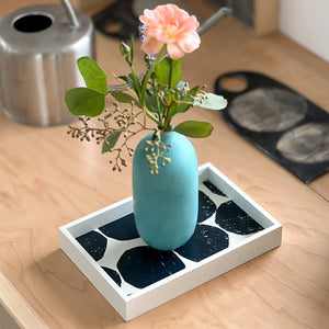 PilgrimWaters Blueberry colorway handcrafted wooden Mini-Mini tray featuring a bold dark blue and white graphic pattern, styled on a wooden tabletop with a turquoise ceramic bud vase holding a pink flower.