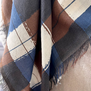 A close-up of a printed plaid scarf with fringed edges, featuring a pattern of blue, brown, cream, and dark gray.