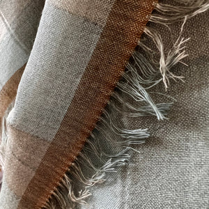 Close-up of a printed textile with a plaid pattern in taupe, brown, and light gray, finished with fringed edges.