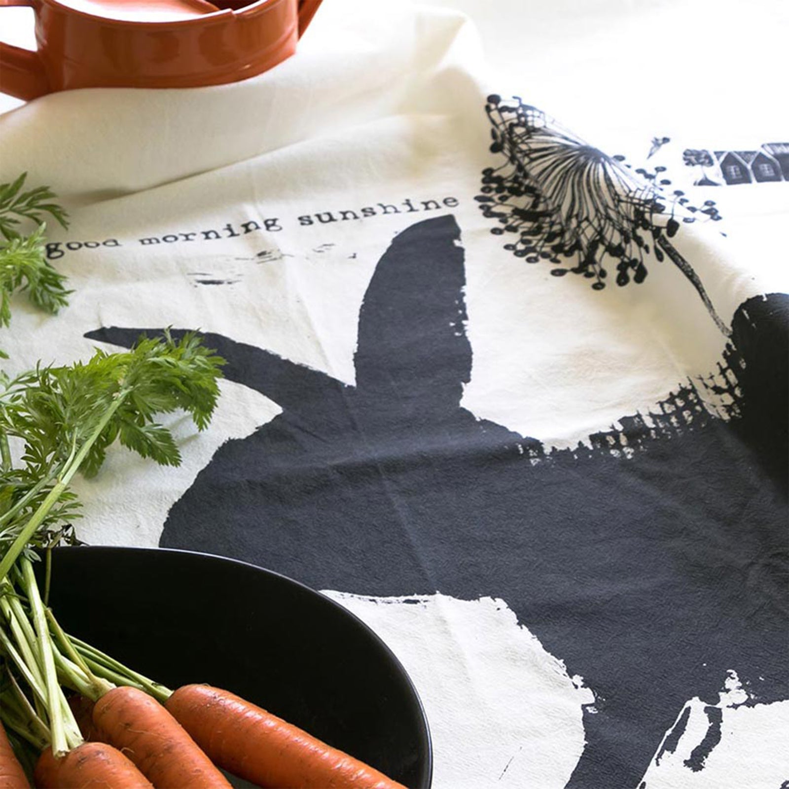Bunny tea towel flat on a surface with carrots and a watering can. Design by Susy Pilgrim Waters. Copyright PilgrimWaters 2026.