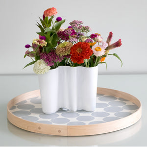 An 18"  round handprinted gray on cotton white with an ash wood band, styled with flowers in a vase in a brightly lit room.