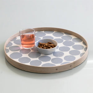 An 18-inch hand-printed round ash-wood gray tray, styled in a brightly lit room with a bowl of nuts and a glass of rose.